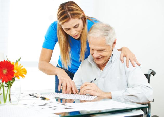 An elderly man in a wheelchair is writing on a piece of paper at an assisted living facility.