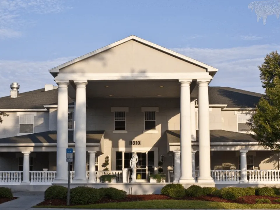 A large, two-story building with white columns and a triangular pediment at the entrance, numbered 1810, serves as one of the premier Florida Assisted Living communities. A white statue and manicured shrubbery enhance its welcoming facade.