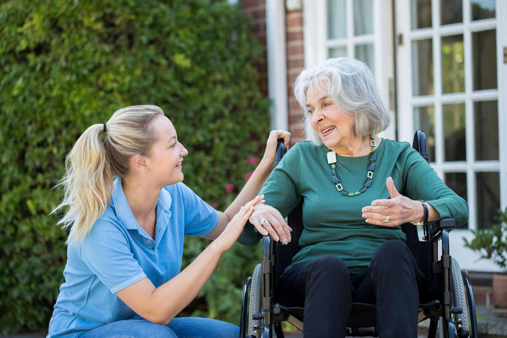 A caregiver in a blue shirt smiles warmly as she interacts with an elderly woman in a wheelchair outside an Ocala, FL assisted living facility near a window.