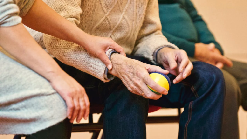 A close-up of an elderly person sitting at Hampton Manor in Ocala, FL, holding a ball, while another person gently touches their arm in the serene setting of assisted living.