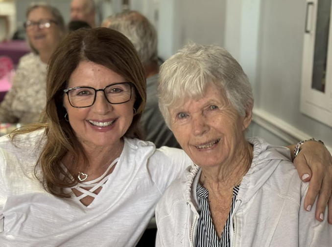Two women, one older and one younger, sit closely together indoors in an assisted living setting, smiling at the camera. The younger woman has glasses and brown hair; the older woman has short gray hair.