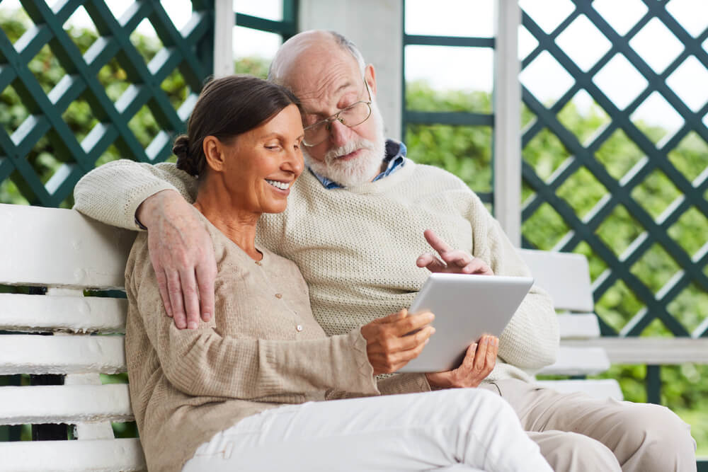 An older couple sits on a white bench outdoors, smiling and looking at a tablet together as they research how to pay for memory care in Florida.