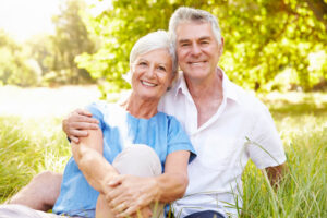 An older couple sits closely together on grass in a sunny park, smiling at the camera. Trees and greenery are visible in the background, reflecting the warmth and tranquility found in some of the best retirement options in Ocala.