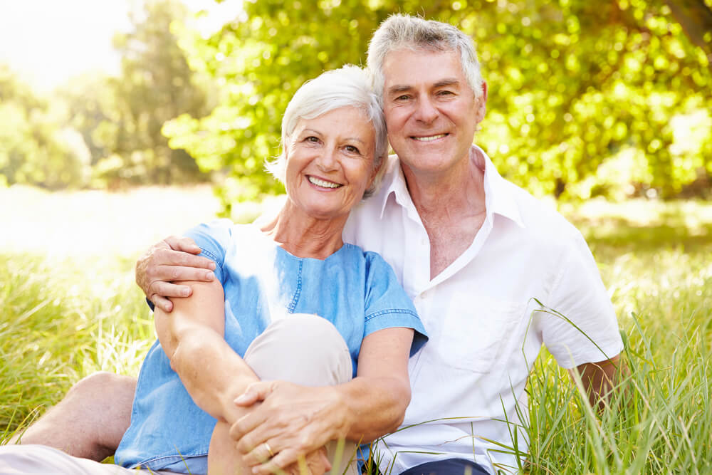 An older couple sits closely together on grass in a sunny park, smiling at the camera. Trees and greenery are visible in the background, reflecting the warmth and tranquility found in some of the best retirement options in Ocala.